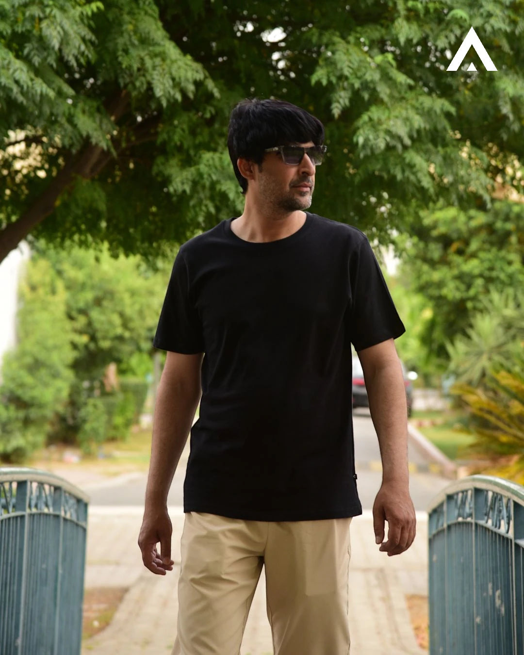 Man wearing a black t-shirt and sunglasses standing outdoors with greenery in the background