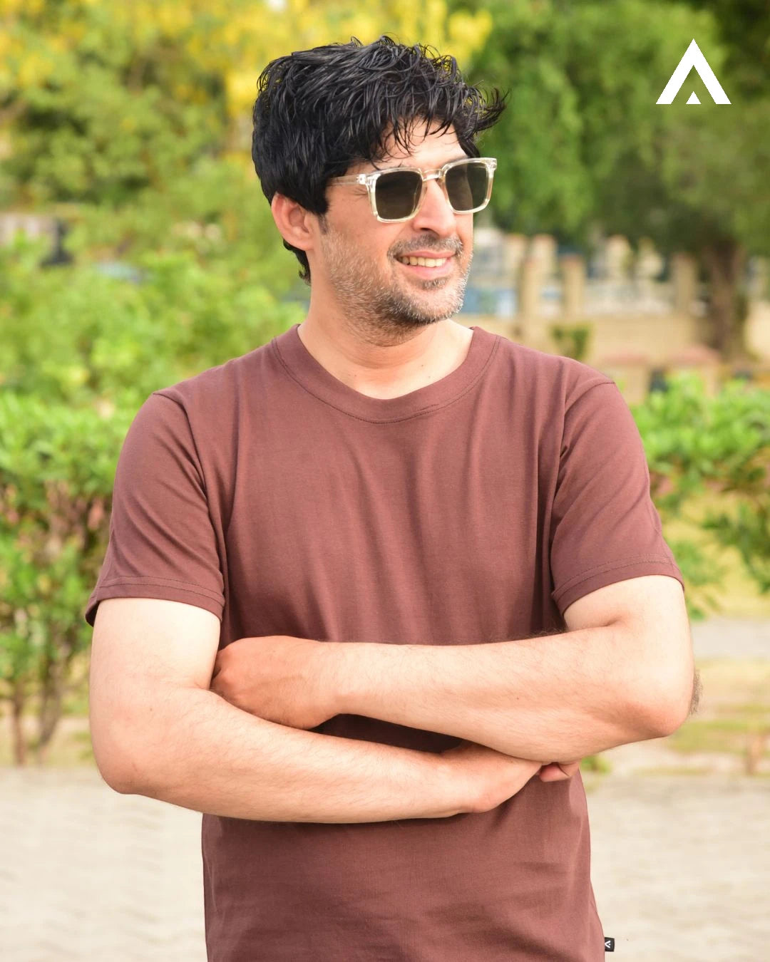 Man wearing sunglasses and a brown t-shirt with a blurred green outdoor background