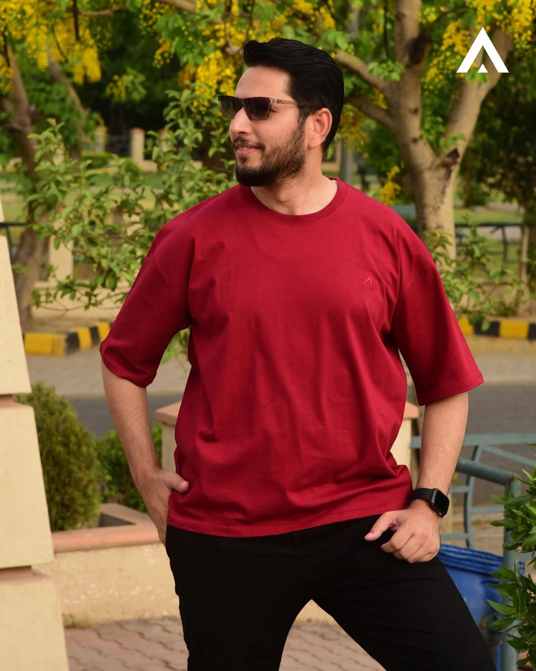 Man wearing a red t-shirt and black pants standing outdoors with trees and a path in the background.