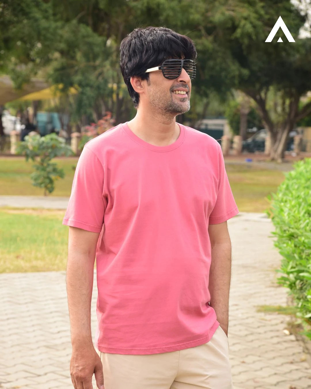 Man wearing a pink shirt and sunglasses outdoors with greenery in the background