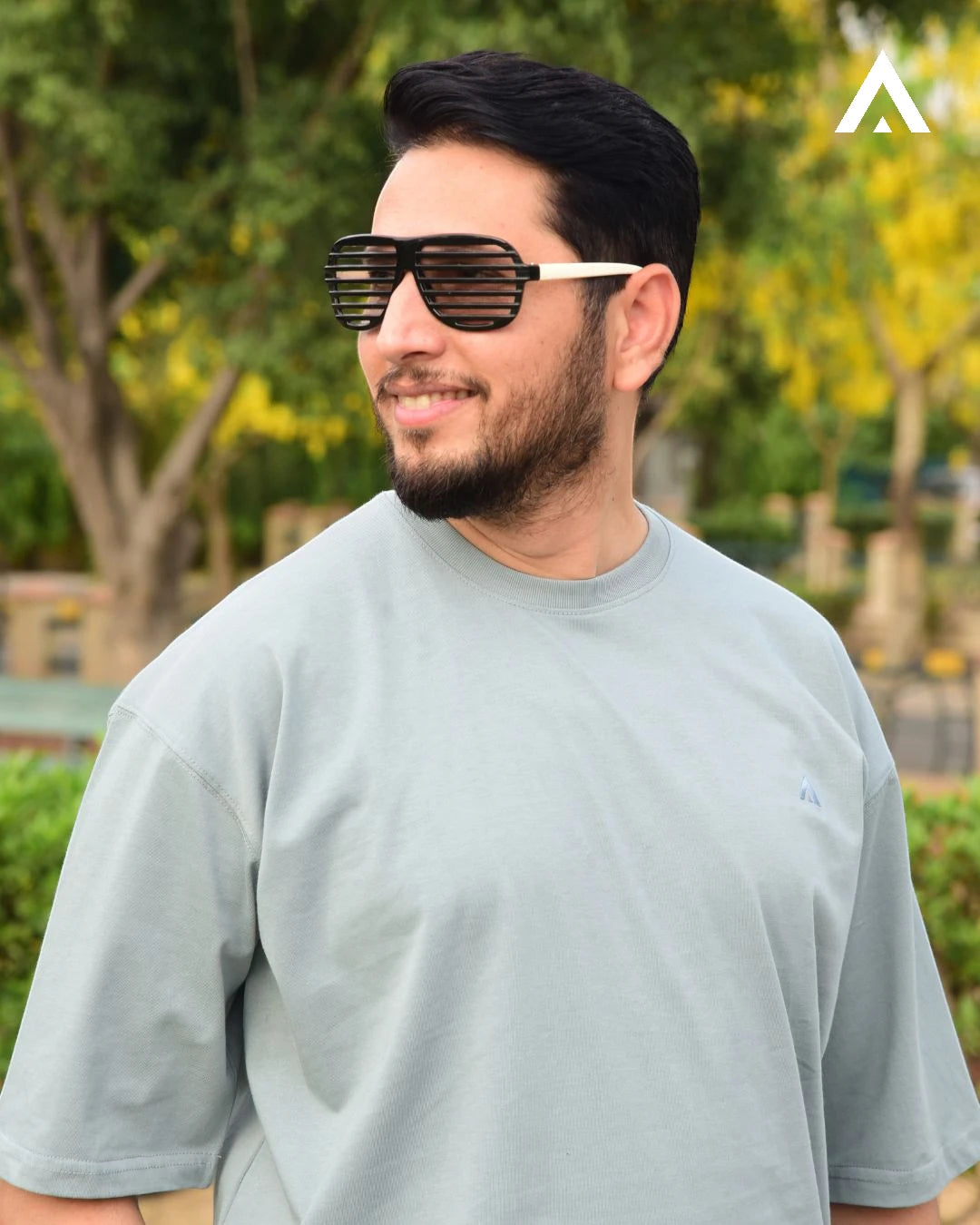 Man wearing sunglasses and a gray t-shirt outdoors with greenery in the background