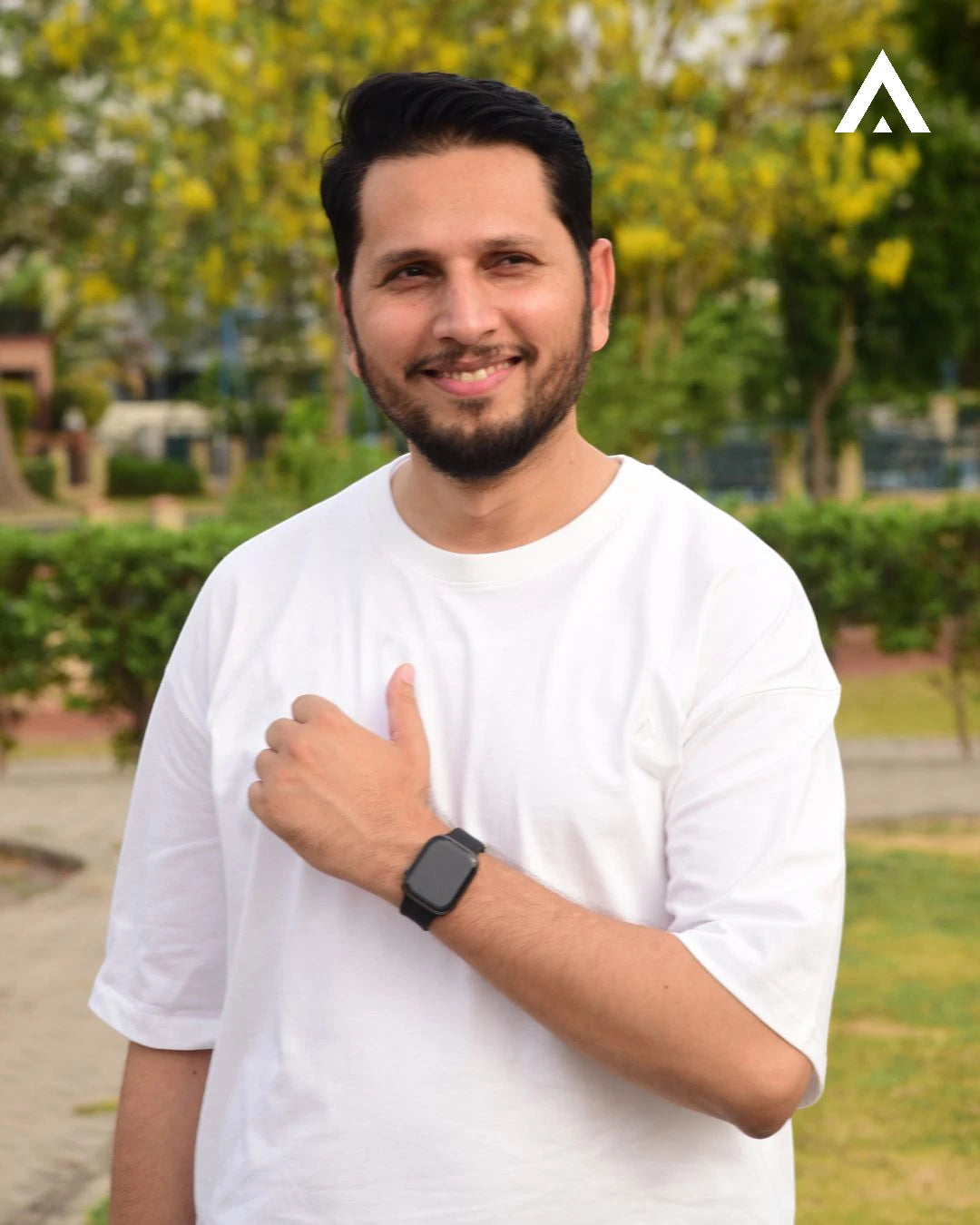 Man wearing a white t-shirt and black watch standing outdoors with greenery in the background